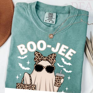 May include: A teal Comfort Colors t-shirt featuring a ghost graphic wearing sunglasses and a leopard print bow, carrying a leopard print bag and coffee cup. The text "BOO-JEE" is above the ghost. Bats and stars surround the ghost.