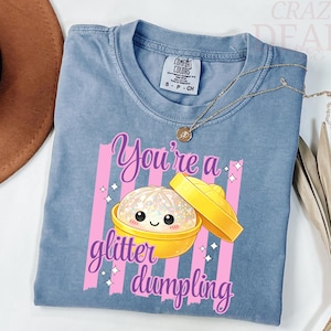 May include: A blue Comfort Colors t-shirt with the text "You're a glitter dumpling" in pink script. The graphic features a cute dumpling with glitter and a yellow lid. A gold necklace is draped across the shirt.