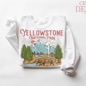 May include: White long-sleeved sweatshirt featuring a Yellowstone National Park graphic. The design includes a bison, mountains, trees, and the text "Yellowstone National Park" in red and black.