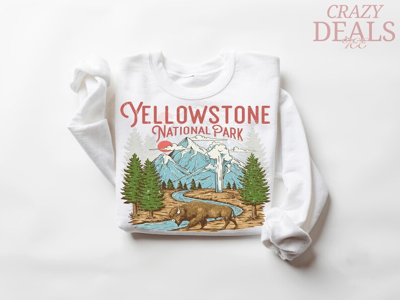 May include: White long-sleeved sweatshirt featuring a Yellowstone National Park graphic. The design includes a bison, mountains, trees, and the text "Yellowstone National Park" in red and black.