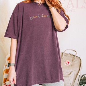 May include: A plum-colored oversized t-shirt with the words "Speech therapy" embroidered in a rainbow thread across the chest. The shirt is paired with gray and white striped shorts. A watch is visible on the left wrist.
