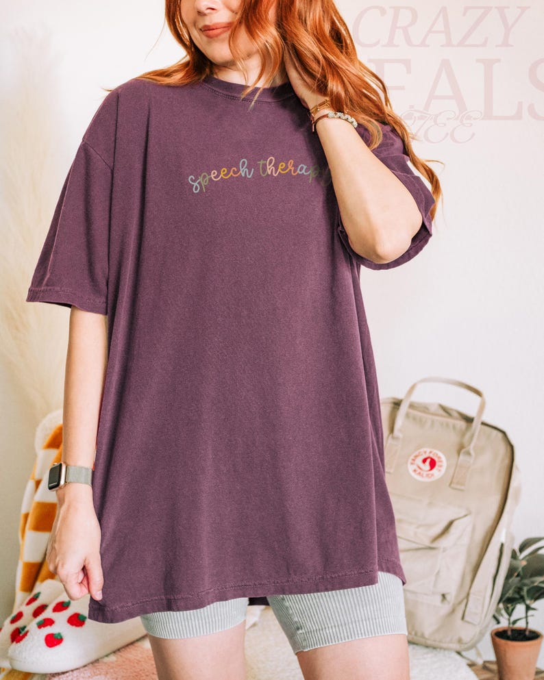 May include: A plum-colored oversized t-shirt with the words "Speech therapy" embroidered in a rainbow thread across the chest. The shirt is paired with gray and white striped shorts. A watch is visible on the left wrist.