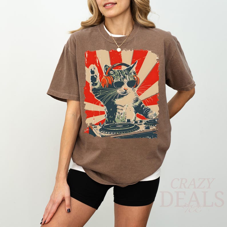 May include: Brown t-shirt with a graphic of a cat DJ wearing sunglasses and headphones, set against a red and cream sunburst design. The cat is behind a turntable. The shirt has a relaxed fit.
