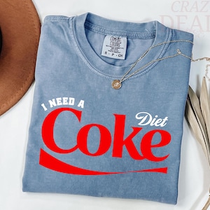 Puede incluir: Una camiseta azul Comfort Colors con las palabras "I NEED A Coke Diet" en rojo y blanco. Un collar de oro con un colgante está drapeado sobre la camisa. La camisa está doblada sobre una superficie blanca.