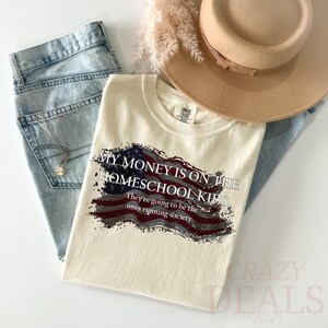 Pu&ograve; includere: T-shirt color crema con la scritta "MY MONEY IS ON THE HOMESCHOOL KIDS" in bianco, con un disegno della bandiera americana. La maglietta &egrave; abbinata a jeans chiari, una collana e un cappello color cammello. La maglietta dice anche "They're going to be the ones running society."