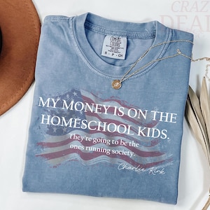 Pu&ograve; includere: Una maglietta blu a maniche corte con la scritta "MY MONEY IS ON THE HOMESCHOOL KIDS." La maglietta presenta un disegno della bandiera americana usurata e la citazione "They're going to be the ones running society." di Charlie Kirk.