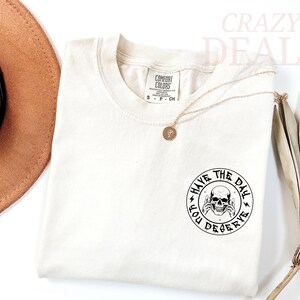 Puede incluir: Camiseta color crema con cuello redondo y mangas cortas. La camiseta presenta un gr&aacute;fico circular con una calavera y el texto "HAVE THE DAY YOU DESERVE". Tambi&eacute;n se ven un collar dorado y un sombrero marr&oacute;n.