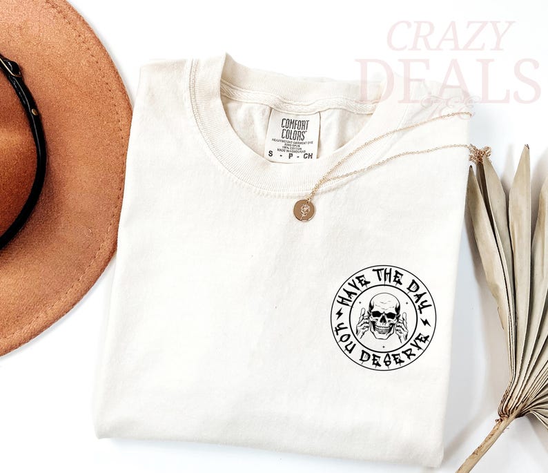 Puede incluir: Camiseta color crema con cuello redondo y mangas cortas. La camiseta presenta un gr&aacute;fico circular con una calavera y el texto "HAVE THE DAY YOU DESERVE". Tambi&eacute;n se ven un collar dorado y un sombrero marr&oacute;n.