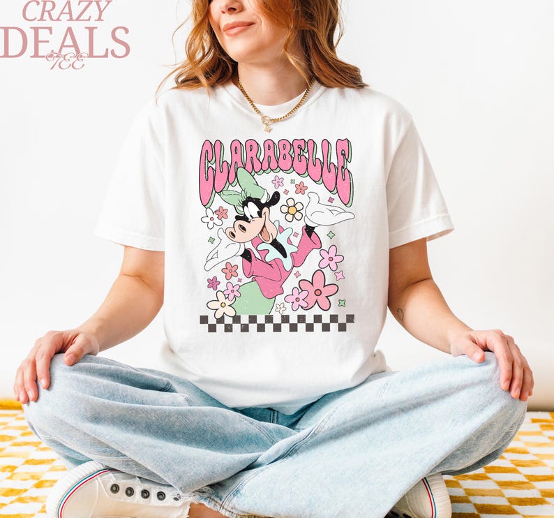 May include: White t-shirt with a cartoon Clarabelle Cow design in pink and green, surrounded by flowers. The word "CLARABELLE" is in pink bubble letters. The words "CRAZY DEALS" are in the upper left corner.