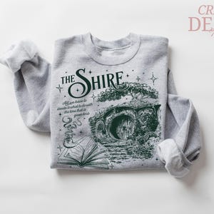 May include: A gray sweatshirt featuring a green graphic of a hobbit hole with the text "THE SHIRE". The design includes a tree, an open book, and a smoking pipe. The text "All we have to decide is what to do with the time that is given us" is also included.