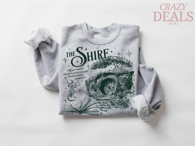 May include: A gray sweatshirt featuring a green graphic of a hobbit hole with the text "THE SHIRE". The design includes a tree, an open book, and a smoking pipe. The text "All we have to decide is what to do with the time that is given us" is also included.