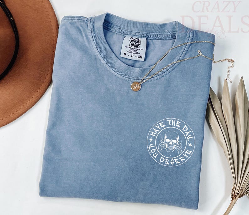 Puede incluir: Una camiseta azul con cuello redondo y mangas cortas. La camiseta tiene un gr&aacute;fico blanco en la espalda que dice "Have the Day You Deserve" con una ilustraci&oacute;n de calavera. Un collar dorado con un colgante est&aacute; sobre la camiseta.
