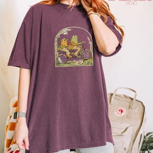 May include: A plum-colored t-shirt featuring a whimsical illustration of two frogs riding a bicycle. The graphic is framed by an archway of foliage. The text "CRAZY DEALS" is printed in pink script at the top right.