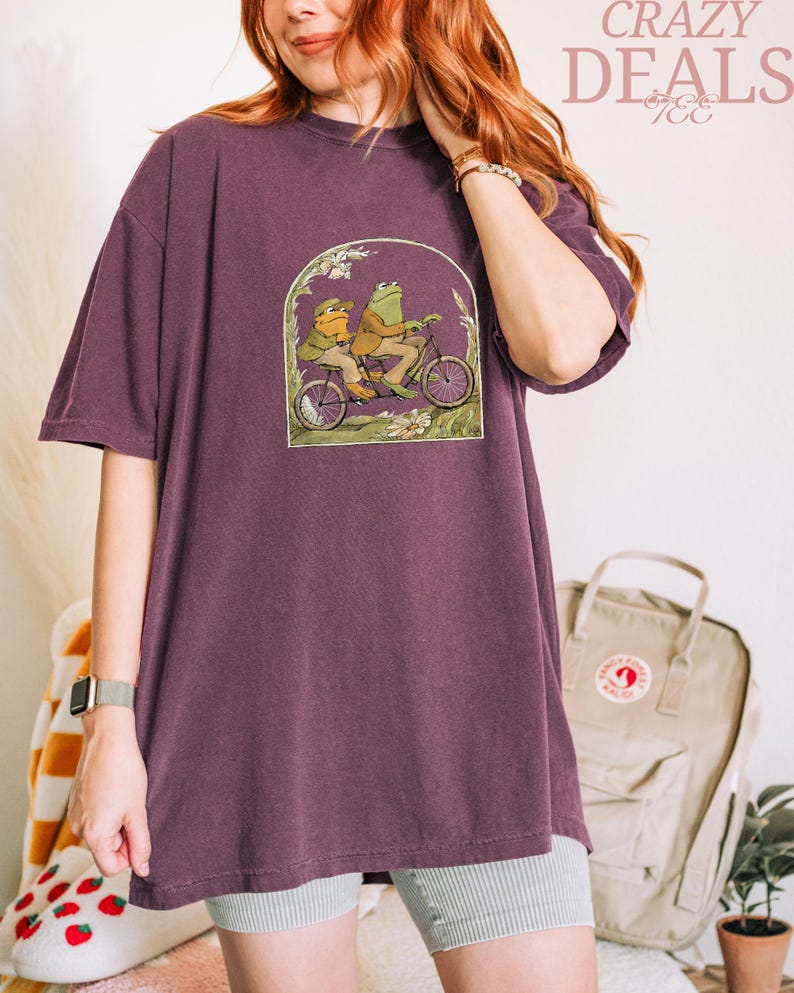 May include: A plum-colored t-shirt featuring a whimsical illustration of two frogs riding a bicycle. The graphic is framed by an archway of foliage. The text "CRAZY DEALS" is printed in pink script at the top right.
