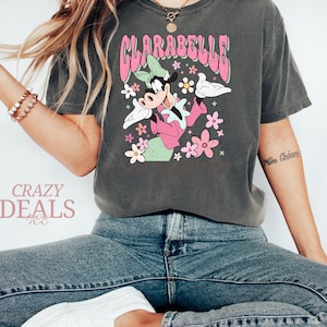 May include: Dark grey t-shirt with a retro Clarabelle Cow graphic, pink text, and flowers. The shirt has a vintage, distressed appearance. The text "Crazy Deals" is visible in the lower left corner.