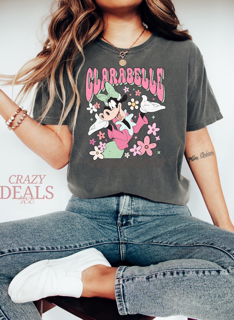 May include: Dark grey t-shirt with a retro Clarabelle Cow graphic, pink text, and flowers. The shirt has a vintage, distressed appearance. The text "Crazy Deals" is visible in the lower left corner.