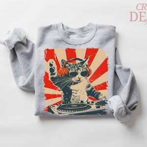 May include: A grey sweatshirt with a graphic of a cat wearing sunglasses and headphones, operating a DJ turntable. The design has a retro style with red and beige accents. The text "CRAZY DEALS" is in the upper right corner.