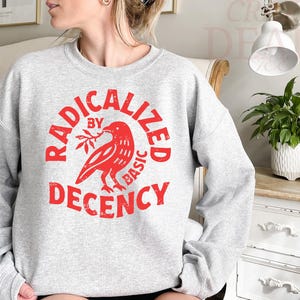 May include: A light gray sweatshirt with a red graphic. The graphic features a bird holding a sprig and the words "RADICALIZED BY BASIC DECENCY" in a circular design. The sweatshirt has long sleeves and a crew neck.