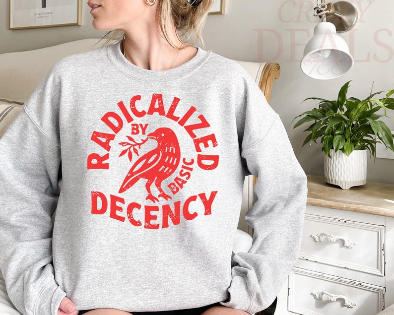 May include: A light gray sweatshirt with a red graphic. The graphic features a bird holding a sprig and the words "RADICALIZED BY BASIC DECENCY" in a circular design. The sweatshirt has long sleeves and a crew neck.