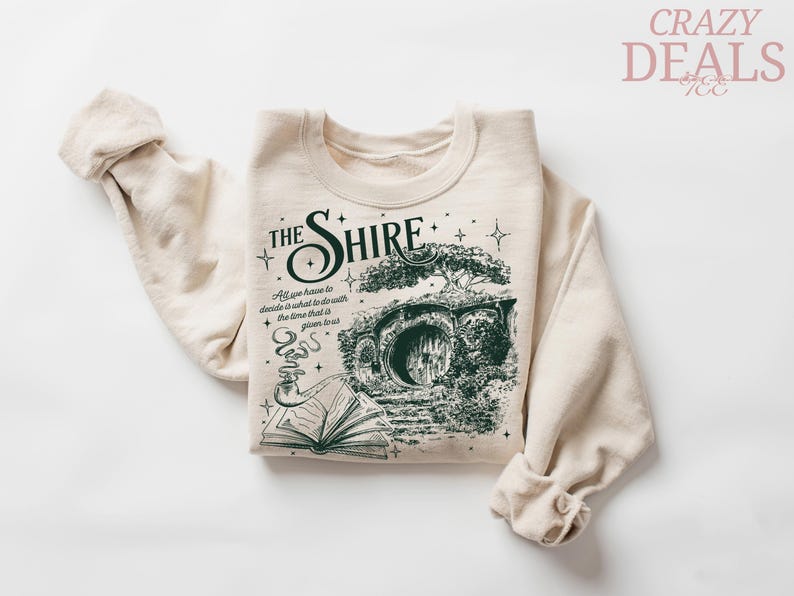 May include: Cream-colored sweatshirt featuring a green graphic of a hobbit hole with the text "The Shire." The design includes an open book and a smoking pipe. The sleeves are rolled up.