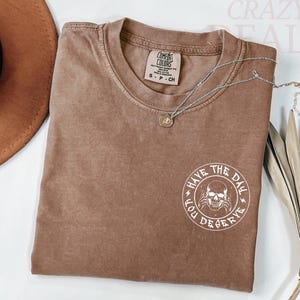 Puede incluir: Una camiseta marr&oacute;n Comfort Colors con un gr&aacute;fico redondo que dice "Have the Day You Deserve" y un dise&ntilde;o de calavera. Un collar plateado con un peque&ntilde;o colgante est&aacute; sobre la camiseta.