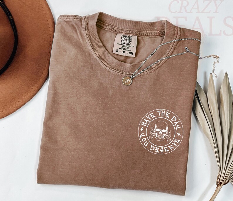 Puede incluir: Una camiseta marr&oacute;n Comfort Colors con un gr&aacute;fico redondo que dice "Have the Day You Deserve" y un dise&ntilde;o de calavera. Un collar plateado con un peque&ntilde;o colgante est&aacute; sobre la camiseta.