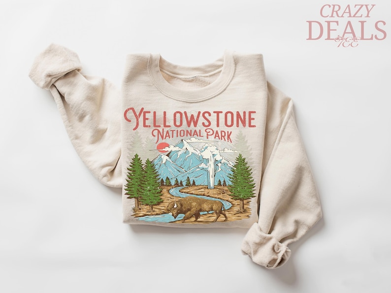 May include: Beige sweatshirt with a graphic of Yellowstone National Park. The design includes mountains, trees, a bison, and the text "Yellowstone National Park" in red. The sleeves are rolled up.