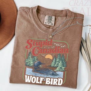 Puede incluir: Camiseta marr&oacute;n con un gr&aacute;fico que dice "Stupid Canadian Wolf Bird" con un colimbo en un lago con &aacute;rboles y una puesta de sol. Un collar plateado con un colgante est&aacute; sobre la camiseta.