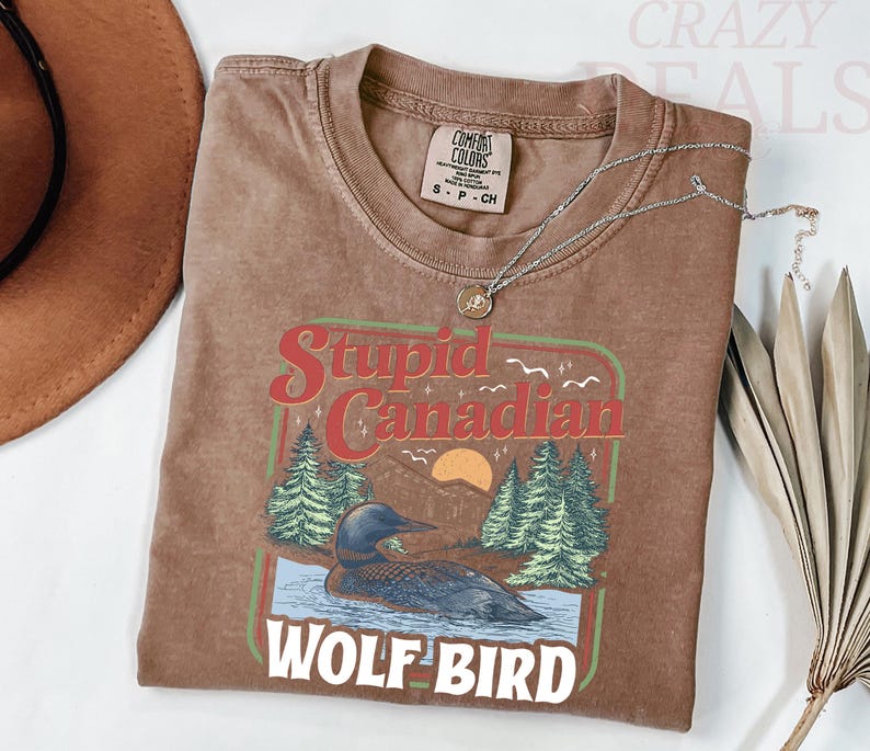 Puede incluir: Camiseta marr&oacute;n con un gr&aacute;fico que dice "Stupid Canadian Wolf Bird" con un colimbo en un lago con &aacute;rboles y una puesta de sol. Un collar plateado con un colgante est&aacute; sobre la camiseta.