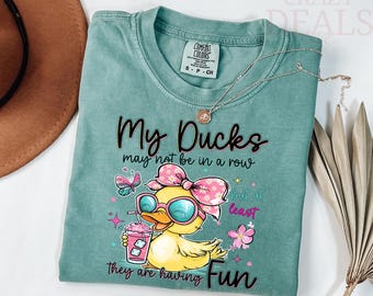 May include: A teal Comfort Colors t-shirt with the text "My Ducks may not be in a row but at least they are having Fun." The shirt features a cartoon duck wearing sunglasses and a pink bow, holding a drink.
