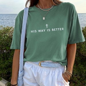 May include: Sage green t-shirt with the text "HIS WAY IS BETTER" and a cross graphic. The shirt is paired with white shorts, a light blue belt, and a matching shoulder bag. Gold jewellery accents the look.