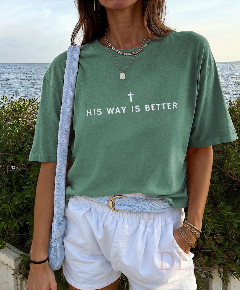 May include: Sage green t-shirt with the text "HIS WAY IS BETTER" and a cross graphic. The shirt is paired with white shorts, a light blue belt, and a matching shoulder bag. Gold jewellery accents the look.