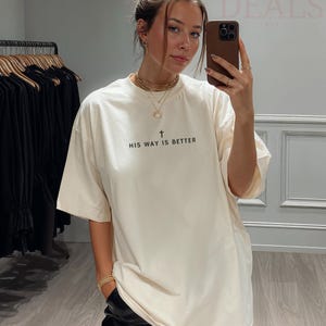 May include: Cream-coloured t-shirt with the text "HIS WAY IS BETTER" in black, featuring a cross symbol. The shirt is a relaxed fit. The image also shows a person wearing the shirt.