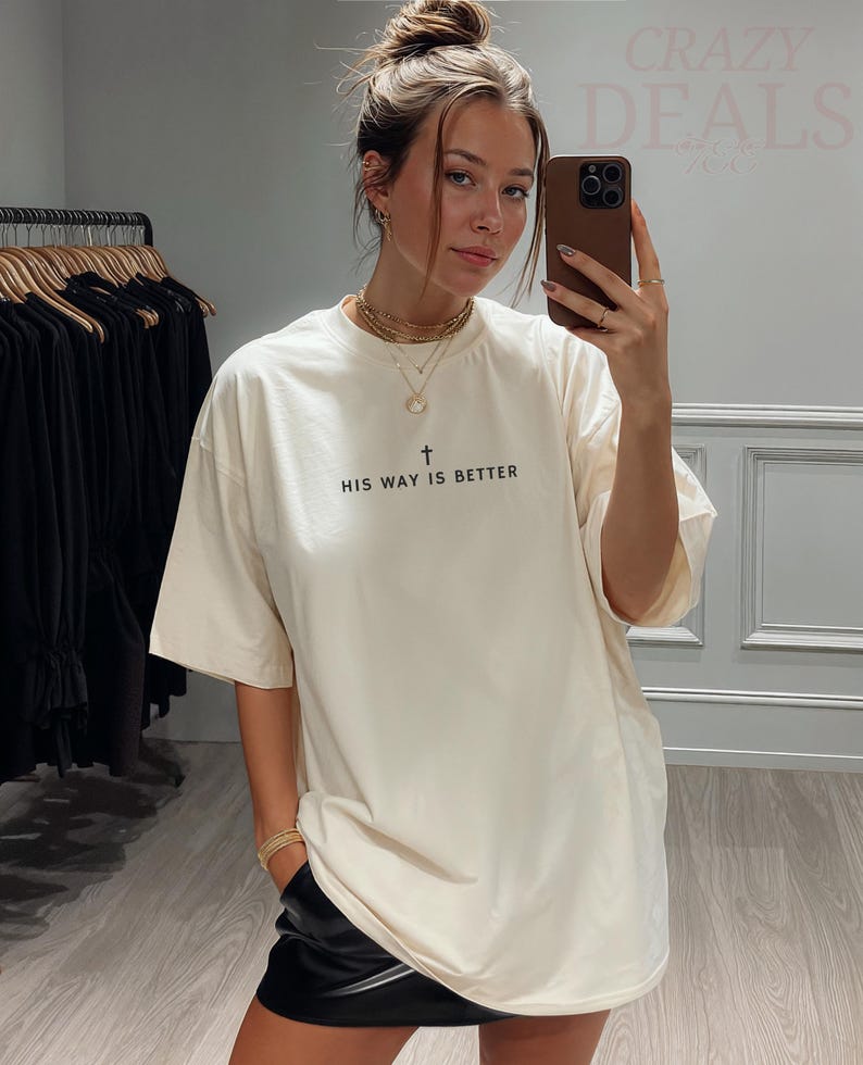 May include: Cream-coloured t-shirt with the text "HIS WAY IS BETTER" in black, featuring a cross symbol. The shirt is a relaxed fit. The image also shows a person wearing the shirt.