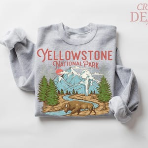 May include: Gray sweatshirt with a graphic of Yellowstone National Park. The design includes mountains, trees, a bison, and the text "Yellowstone National Park" in red. The sweatshirt has rolled-up sleeves.