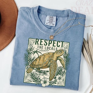 May include: A light blue t-shirt with a graphic of a sea turtle and the text "Respect the Locals." The design includes palm fronds and hibiscus flowers. A gold necklace is draped across the shirt.