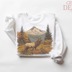 May include: White sweatshirt with a detailed illustration of an elk in a mountain landscape. The design includes a mountain, trees, and autumn foliage. The text "CRAZY DEALS" is in the upper right corner.