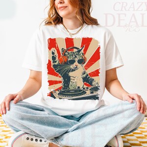 May include: White t-shirt with a graphic of a cat DJ wearing sunglasses and headphones, set against a red and beige retro backdrop. The cat is behind a DJ turntable, with its paw raised. The text "Crazy Deals" is visible.