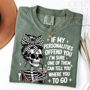 Puede incluir: Camiseta verde oliva con un gráfico de esqueleto con gafas de sol, un lazo a cuadros y una taza de café. La camiseta tiene el texto: "If my personalities offend you, I'm sure one of them can tell you where to go."