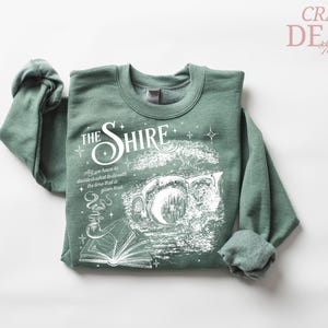May include: Sage green sweatshirt with the text "The Shire" in a stylized font, along with an illustration of a hobbit hole, a book, and a smoking pipe. The sweatshirt also has the quote "All we have to decide is what to do with the time that is given us."