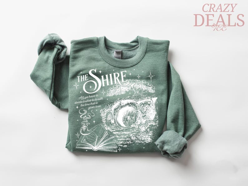 May include: Sage green sweatshirt with the text "The Shire" in a stylized font, along with an illustration of a hobbit hole, a book, and a smoking pipe. The sweatshirt also has the quote "All we have to decide is what to do with the time that is given us."