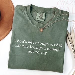 Könnte beinhalten: Olivgrünes T-Shirt mit dem weißen Text "i don't get enough credit for the things i manage not to say". Eine goldene Halskette mit Anhänger befindet sich auf dem T-Shirt. Ein brauner Hut ist im Hintergrund.