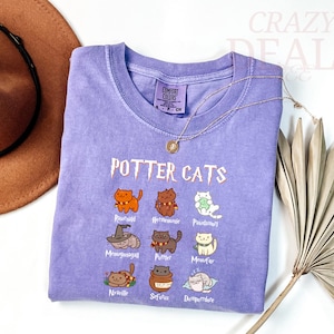 Pu&ograve; includere: Una maglietta lilla con la scritta "POTTER CATS" e illustrazioni di gatti dei cartoni animati. La maglietta presenta vari personaggi di gatti con nomi come "Rawhald" e "Hermetumie". Sono visibili anche una collana d'oro e un cappello marrone.