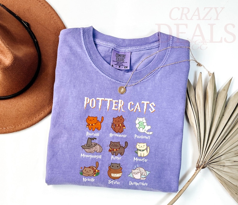 Pu&ograve; includere: Una maglietta lilla con la scritta "POTTER CATS" e illustrazioni di gatti dei cartoni animati. La maglietta presenta vari personaggi di gatti con nomi come "Rawhald" e "Hermetumie". Sono visibili anche una collana d'oro e un cappello marrone.