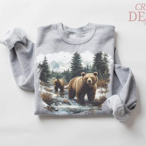 May include: A gray sweatshirt featuring a detailed illustration of two brown bears in a forest stream with evergreen trees and mountains in the background. The text "CRAZY DEALS" is in the upper right corner.