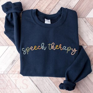 May include: A navy blue sweatshirt with the words "speech therapy" in a colorful, cursive font across the front. The sweatshirt has a crew neck and long sleeves with ribbed cuffs. The garment is made of a soft material.