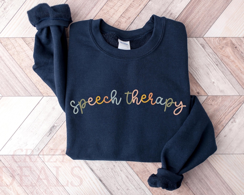 May include: A navy blue sweatshirt with the words "speech therapy" in a colorful, cursive font across the front. The sweatshirt has a crew neck and long sleeves with ribbed cuffs. The garment is made of a soft material.