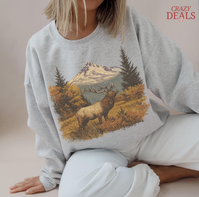 May include: A pale grey sweatshirt with a graphic of an elk in a mountain scene. The design includes a mountain, trees, and autumn colours. The words "CRAZY DEALS" are in the upper right corner.