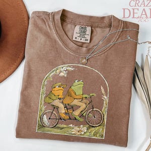 May include: Brown t-shirt with a graphic of two frogs riding a tandem bicycle. The shirt has a crew neck and a small tag at the neckline. A silver necklace with a small pendant is draped across the shirt. The words "Crazy Deals" are in the upper right corner.