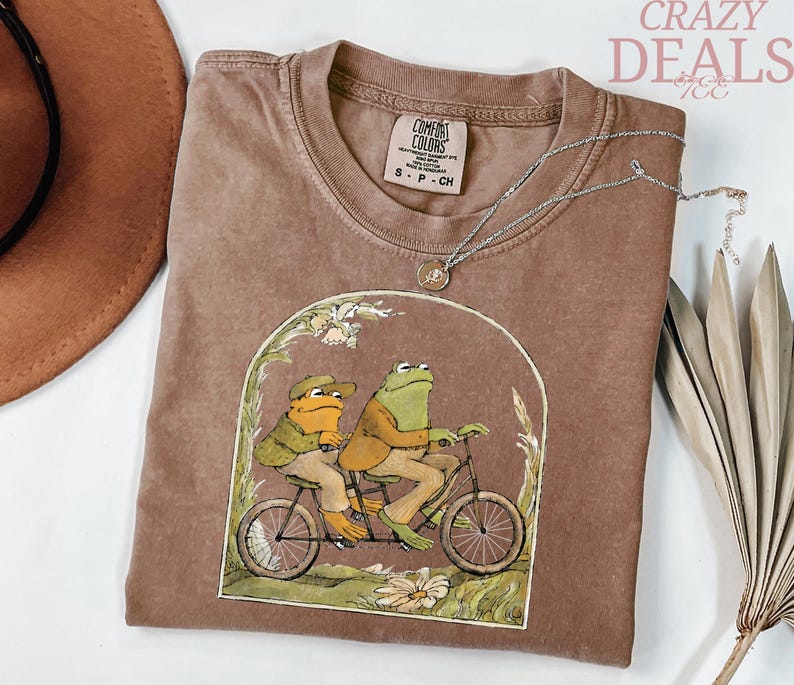 May include: Brown t-shirt with a graphic of two frogs riding a tandem bicycle. The shirt has a crew neck and a small tag at the neckline. A silver necklace with a small pendant is draped across the shirt. The words "Crazy Deals" are in the upper right corner.